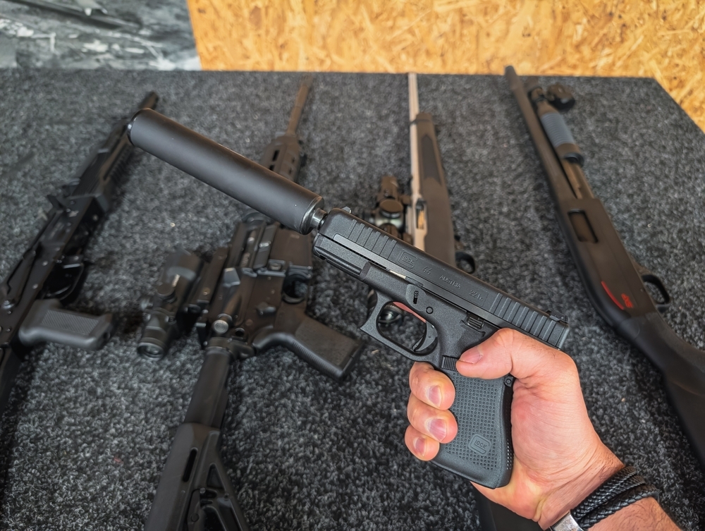 A man holds a Glock 44 pistol with a silencer against a background of other weapons at a shooting range.