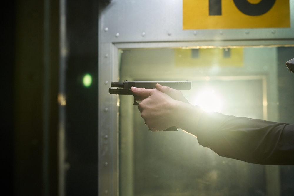 Close-up image showing shooter holding black handgun at indoor gun range. Bright lights reflecting off metal surfaces and number ten visible in background