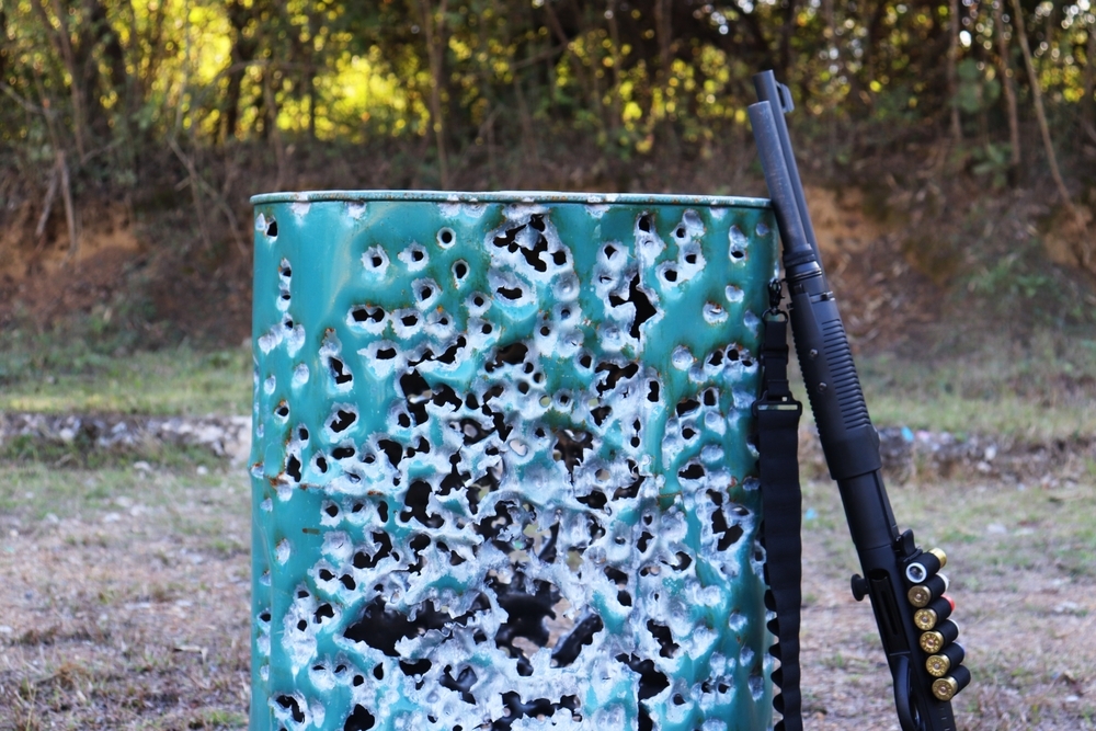 A shotgun rested against the side of a porous green steel barrel. that was shot with dozens of shotgun rounds